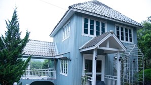 Ein zweistöckiges Haus mit blauer Fassade, einer Veranda mit weißem Geländer und einem kleinen Garten.