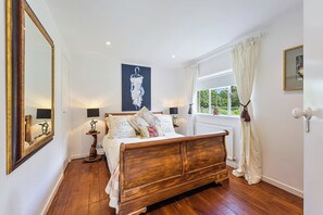 Ein Schlafzimmer mit einem hölzernen Bett, Nachttischlampen, einem großen Spiegel, einem Fenster mit Vorhängen und einem Gemälde an der Wand.