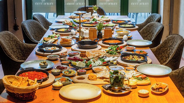 Une table à manger dressée avec différents plats, entourée de chaises, dans une pièce dotée de grandes fenêtres.