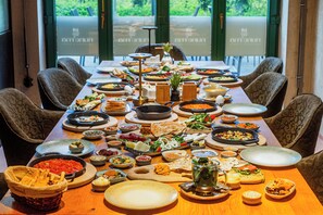 Una mesa de comedor con diversos platos, rodeada de sillas, en una habitación con ventanas grandes.