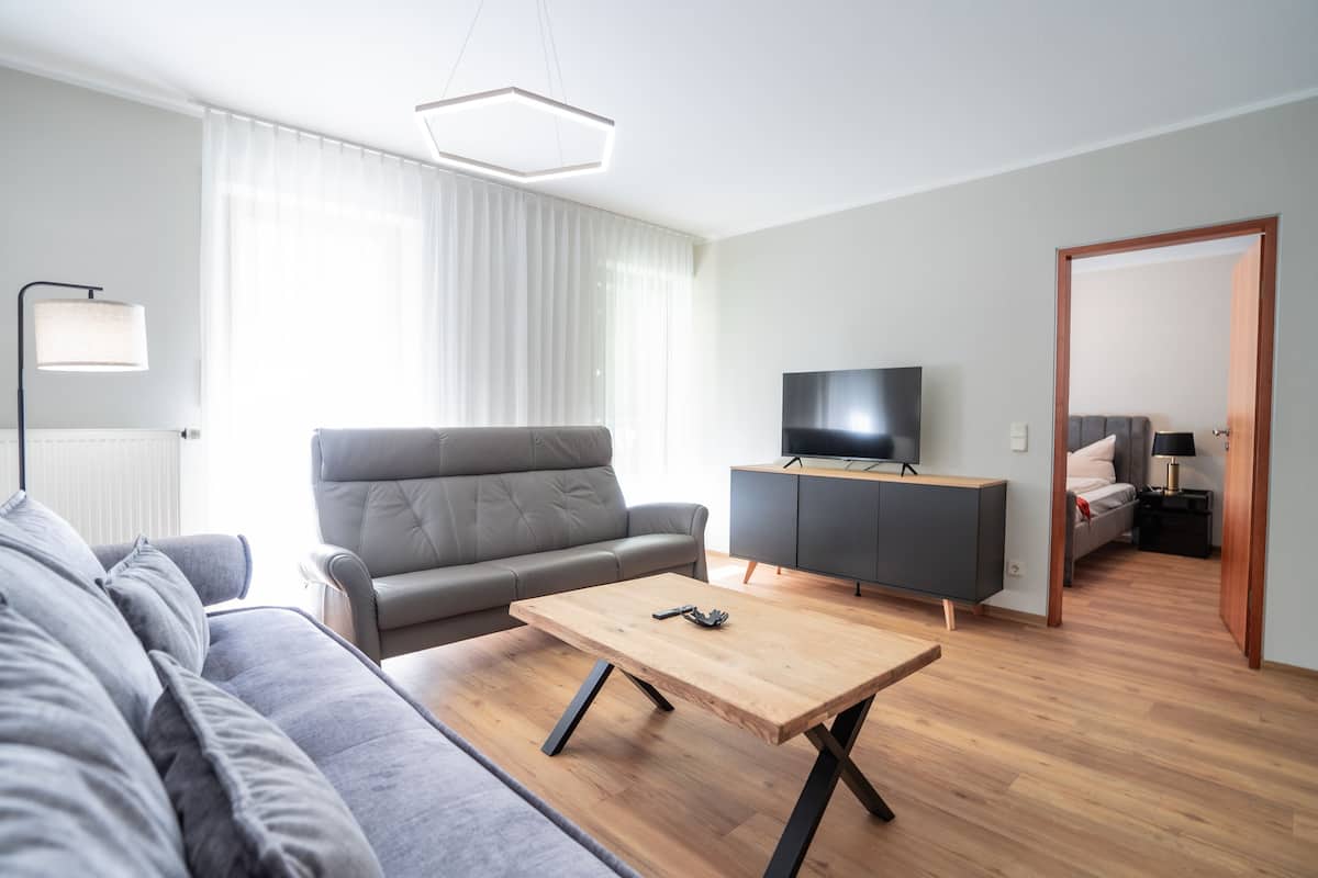 Ein modernes Wohnzimmer mit einem grauen Sofa, einem Holztisch, einem Fernseher auf einem Schrank und einem Flur, der in einen anderen Raum führt.