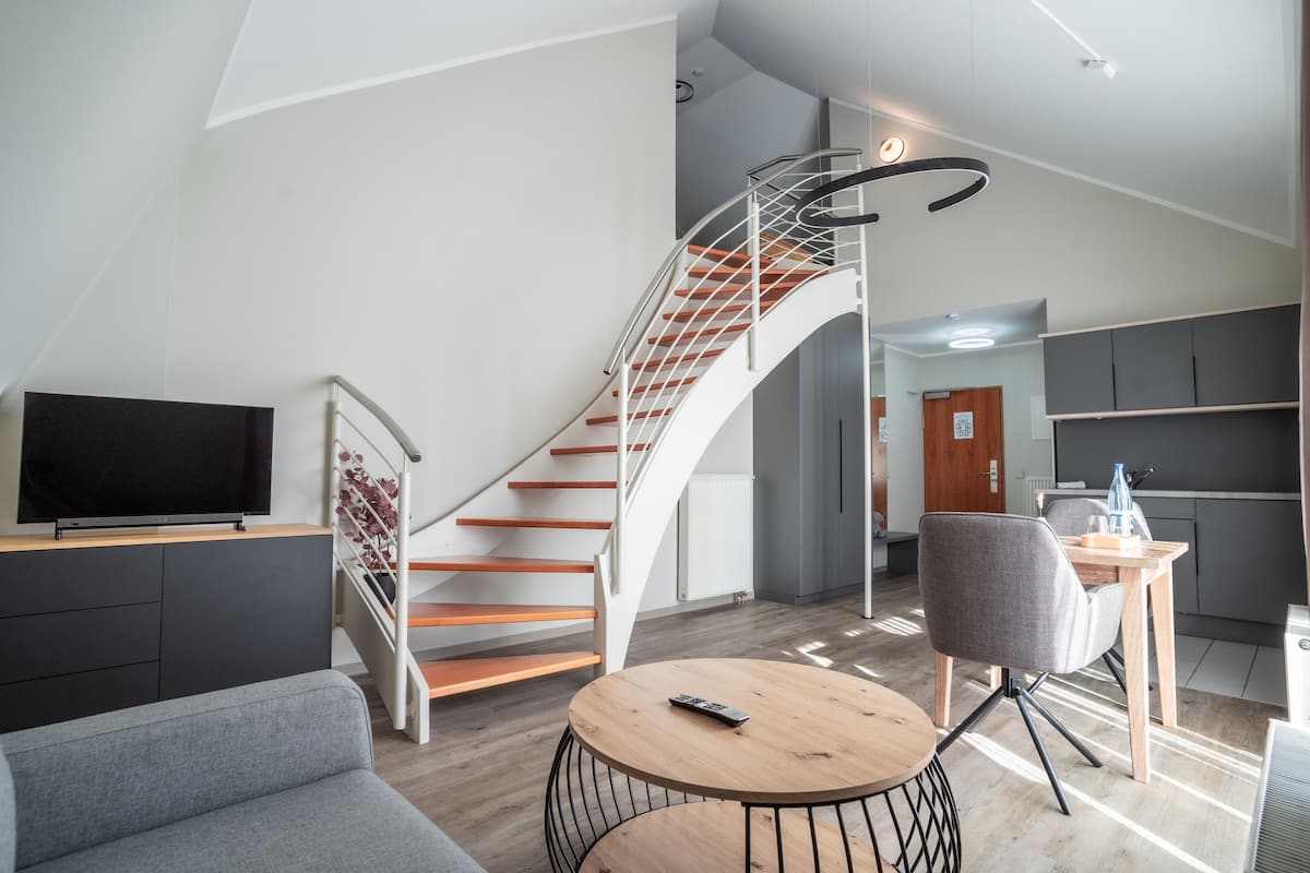 Ein modernes Wohnzimmer mit einer geschwungenen Treppe, einem Flachbildfernseher, einem runden Holztisch und einem grauen Sofa.