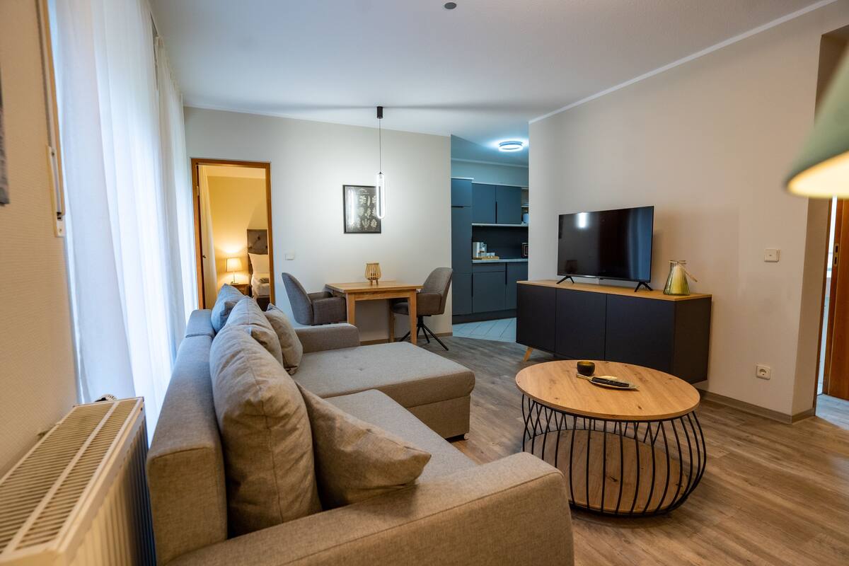 Ein modernes Wohnzimmer mit einer Eckcouch, einem runden Couchtisch, einem Flachbildfernseher und einer Küche im Hintergrund.