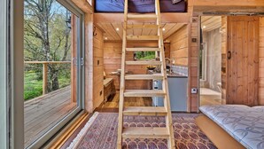 En stuga med träinredning, ett sovloft, en stege, ett köksutrymme och ett stort fönster med utsikt över en skog.