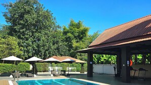 Una piscina con agua cristalina azul, rodeada de sillas de descanso y sombrillas, además de un área de estar cubierta.