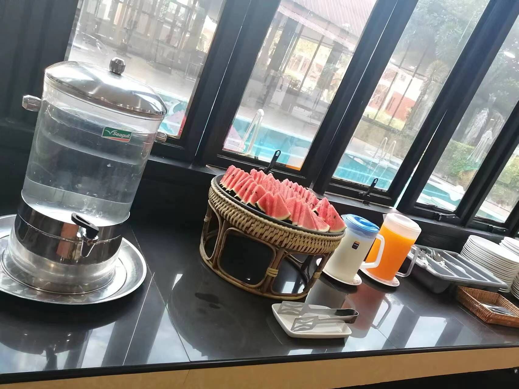 A breakfast setup with a water dispenser, a basket of watermelon slices, a plate with utensils, and a glass of orange juice.