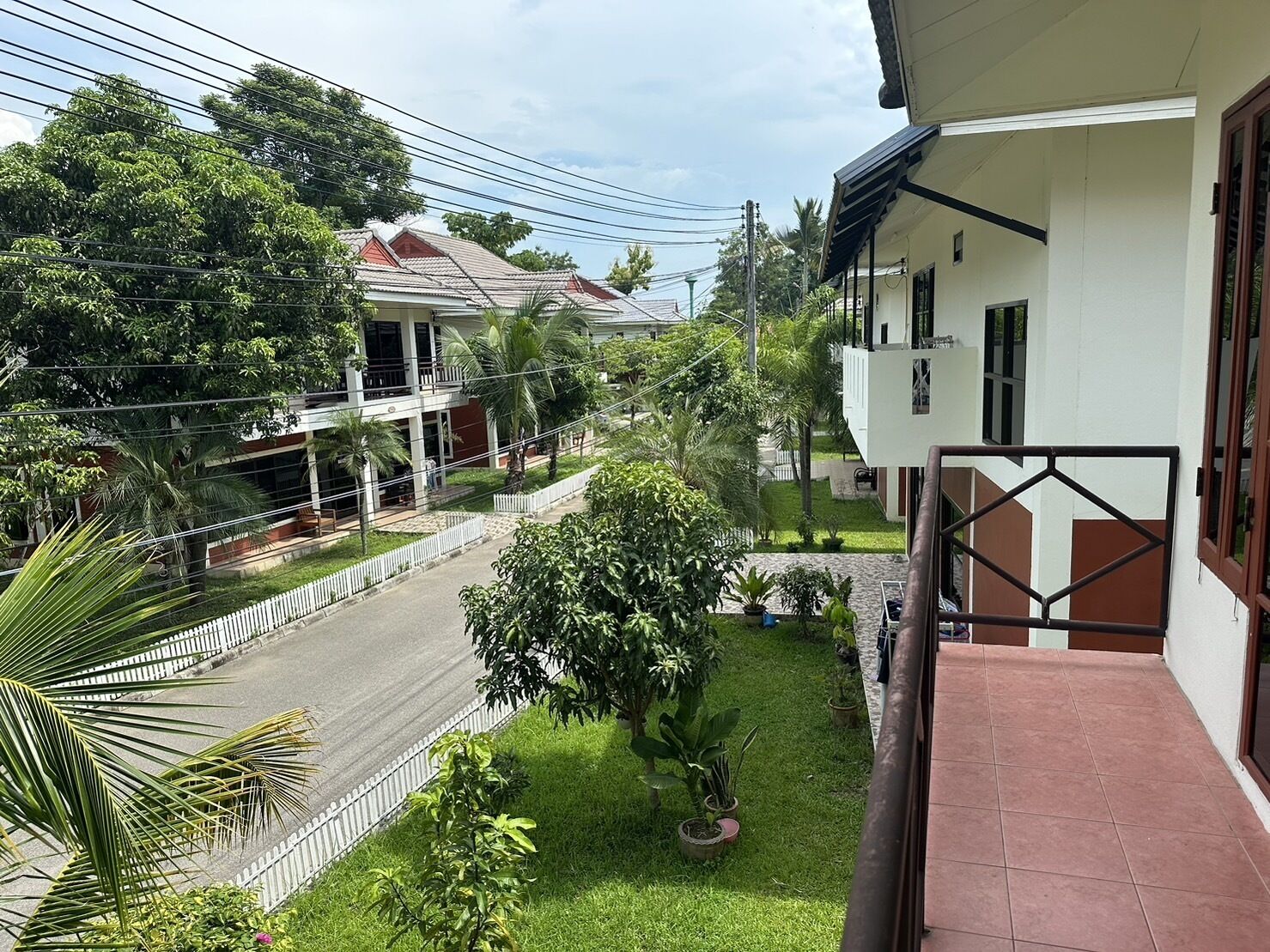 Un barrio residencial con una casa de dos pisos, un balcón y una calle con cerca blanca y follaje.