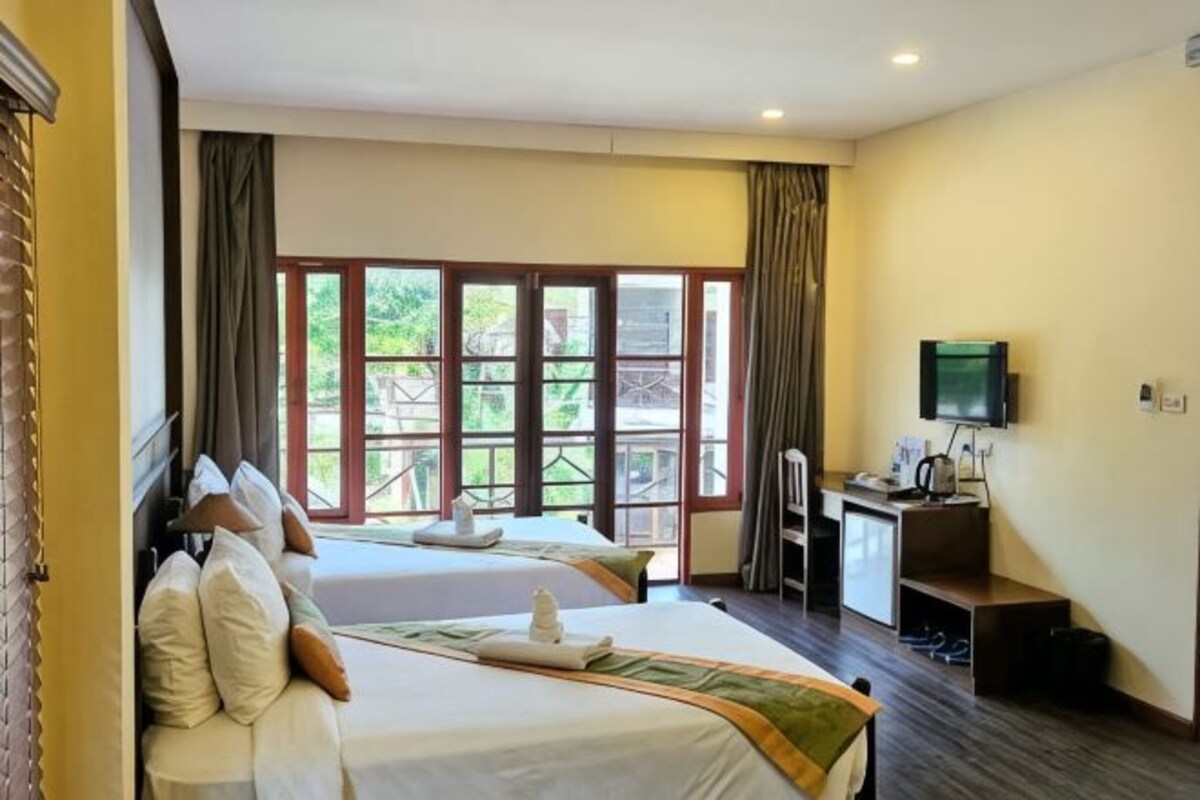 Quarto de hotel com duas camas, uma escrivaninha, televisão e varanda com vista para o verde.