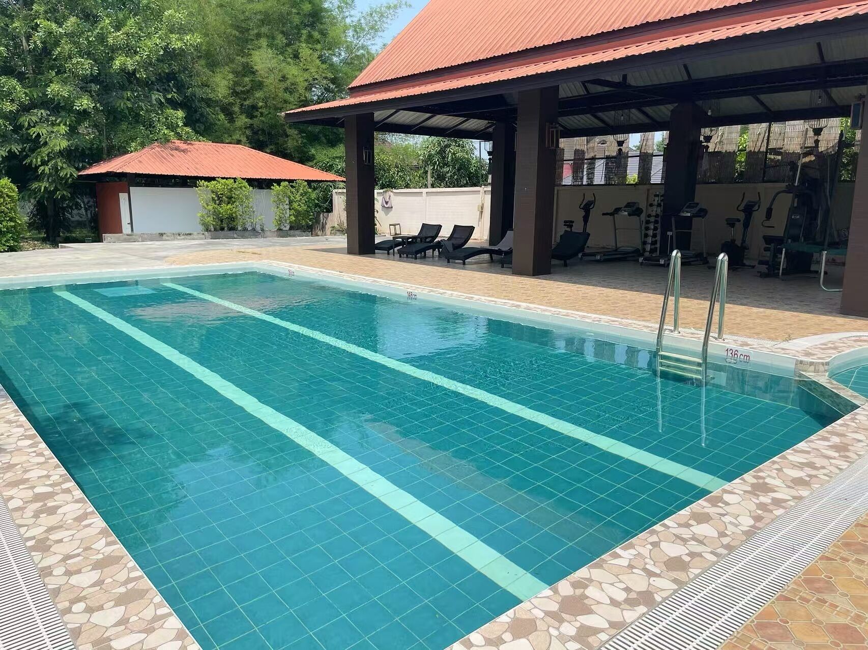 Une piscine rectangulaire à l’eau d’un bleu limpide, dotée d’une plage carrelée et d’un espace abrité équipé de matériel d’exercice.