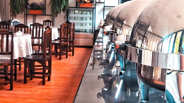 Un restaurante con una barra larga, sillas de madera y mesas. Hay plantas en macetas sobre estantes y una parrilla grande en primer plano.