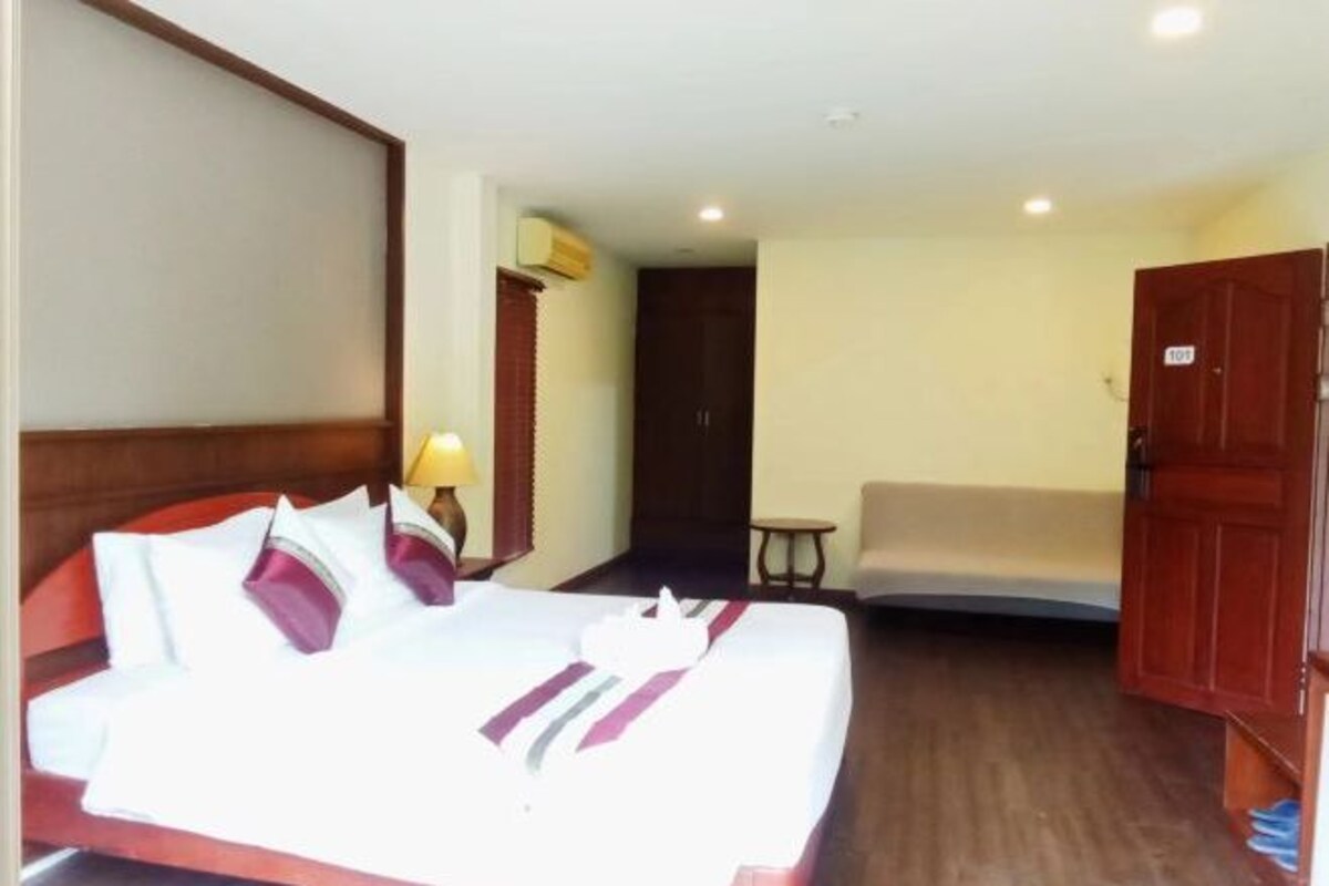 Quarto de hotel com cama bem arrumada, pequena mesa de cabeceira com abajur, porta e uma cadeira.