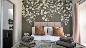Ein ordentlich gemachtes Bett mit einer grauen Strickdecke und zwei braunen Kissen, eine Wand mit Wandmalerei von Blättern und ein dunkler Holzschrank.