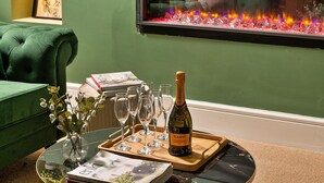 Ein grünes, gepolstertes Sofa, ein Glastisch mit einer Flasche Canti Prosecco, Weingläser und einer Ablagefläche.