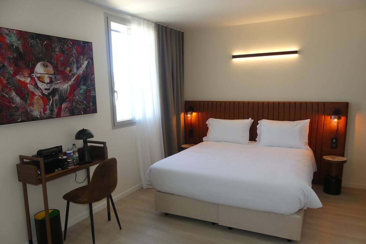 A hotel room with a large bed, a desk with a chair, a painting on the wall, and a window with curtains.