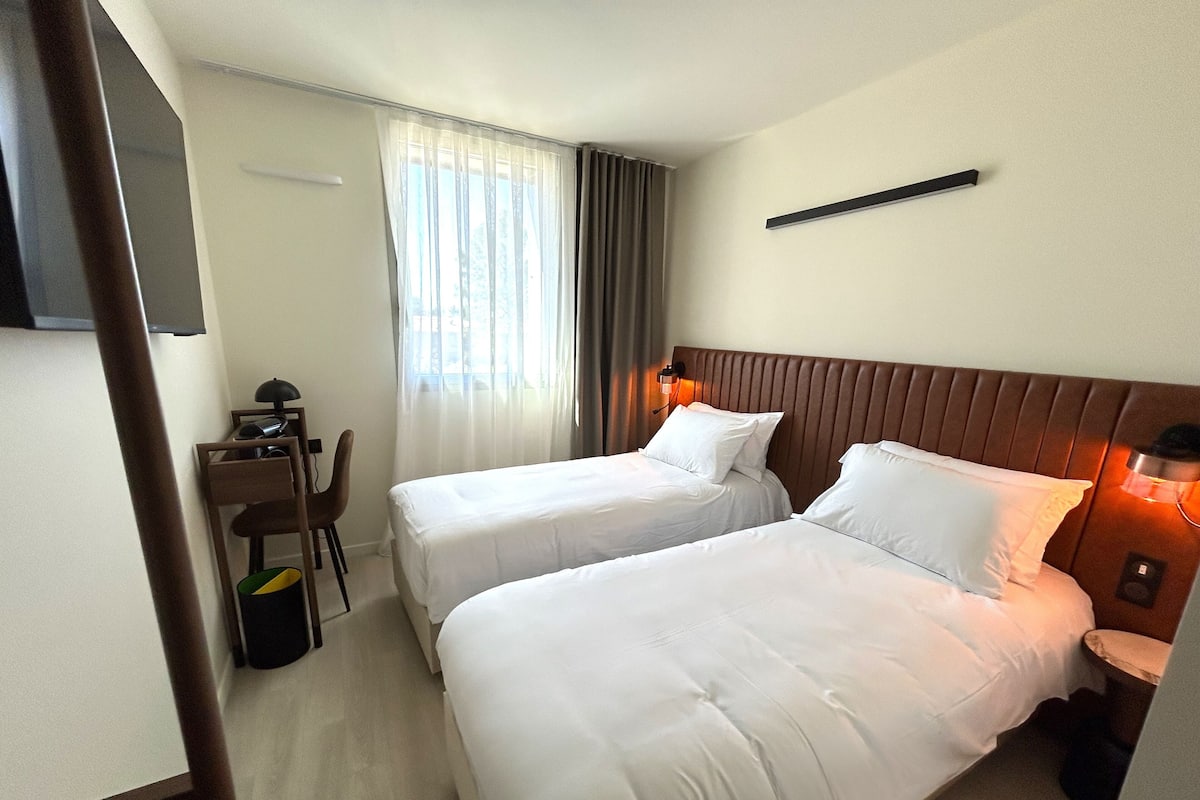 A hotel room with two single beds, a desk with a chair, and a television on the wall.