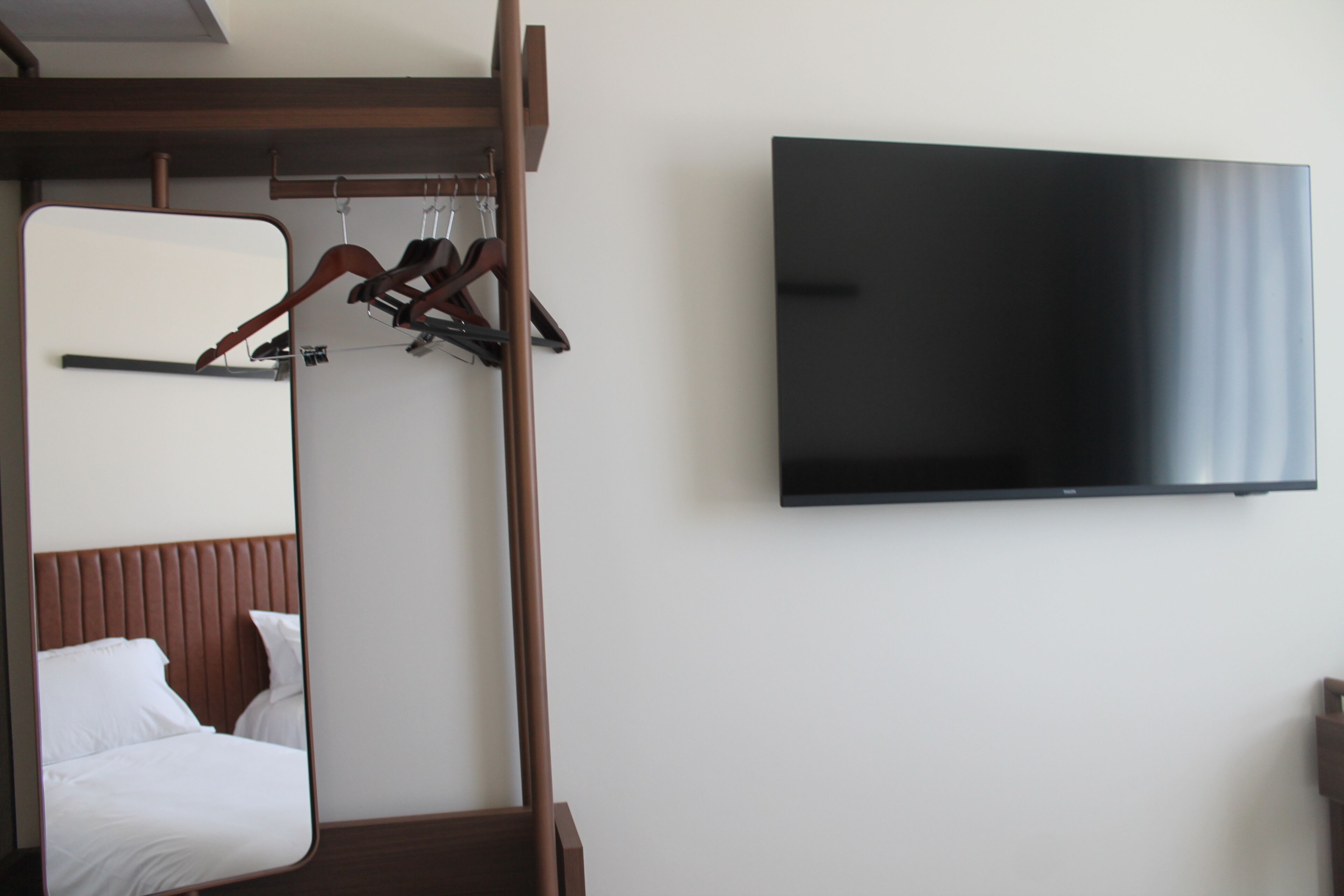 A hotel room with a bed, a mirror, a TV mounted on the wall, and a coat rack with hangers.