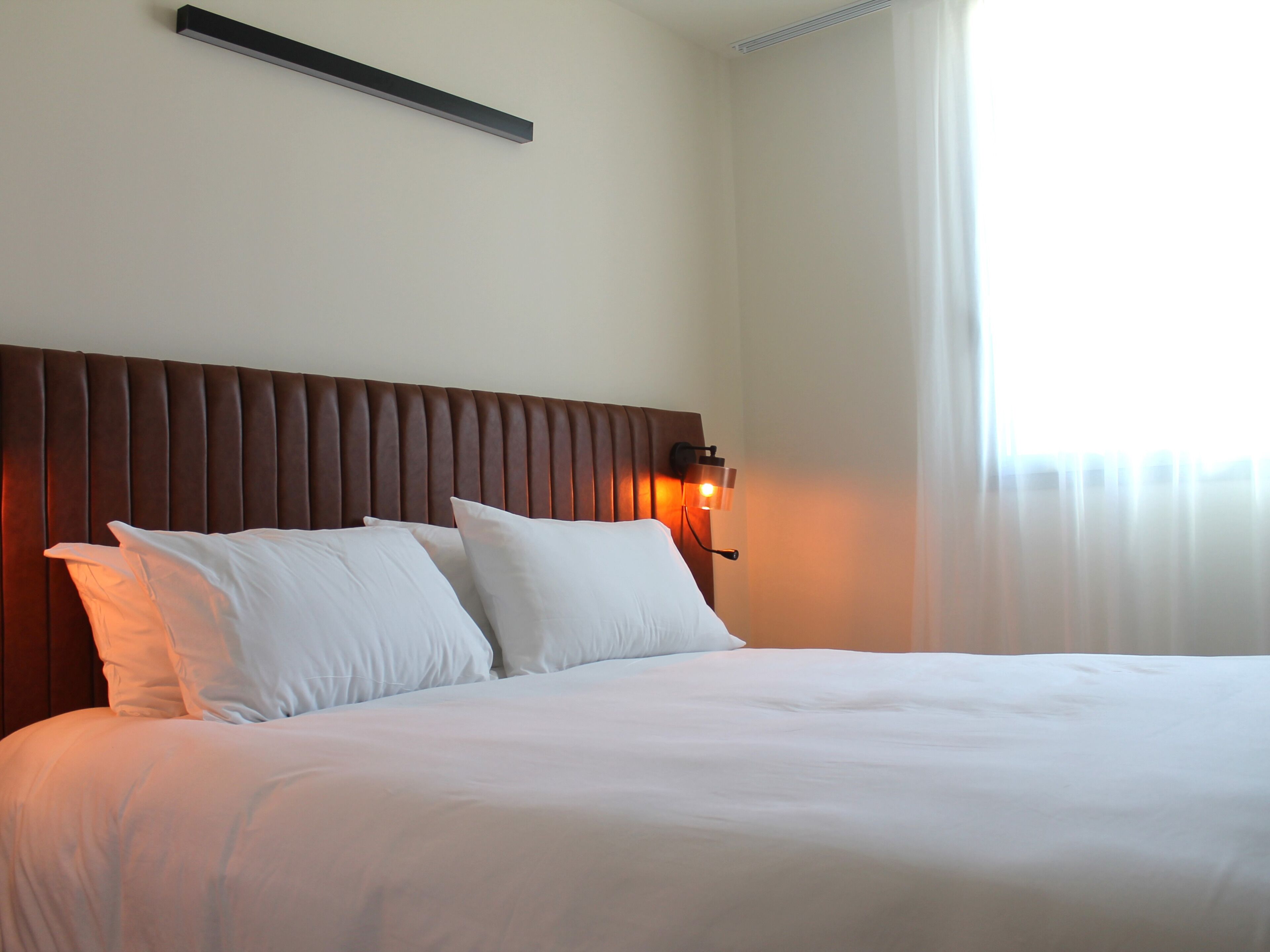 A neatly made bed with white linens and pillows, a wooden headboard, and a wall-mounted light fixture.