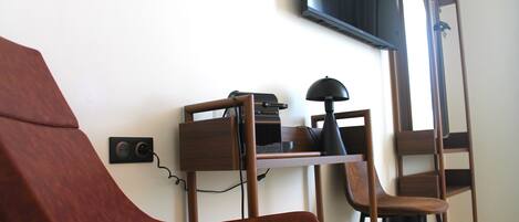 Una habitación con una silla de madera, un escritorio con una computadora, una lámpara y un televisor montado en la pared.