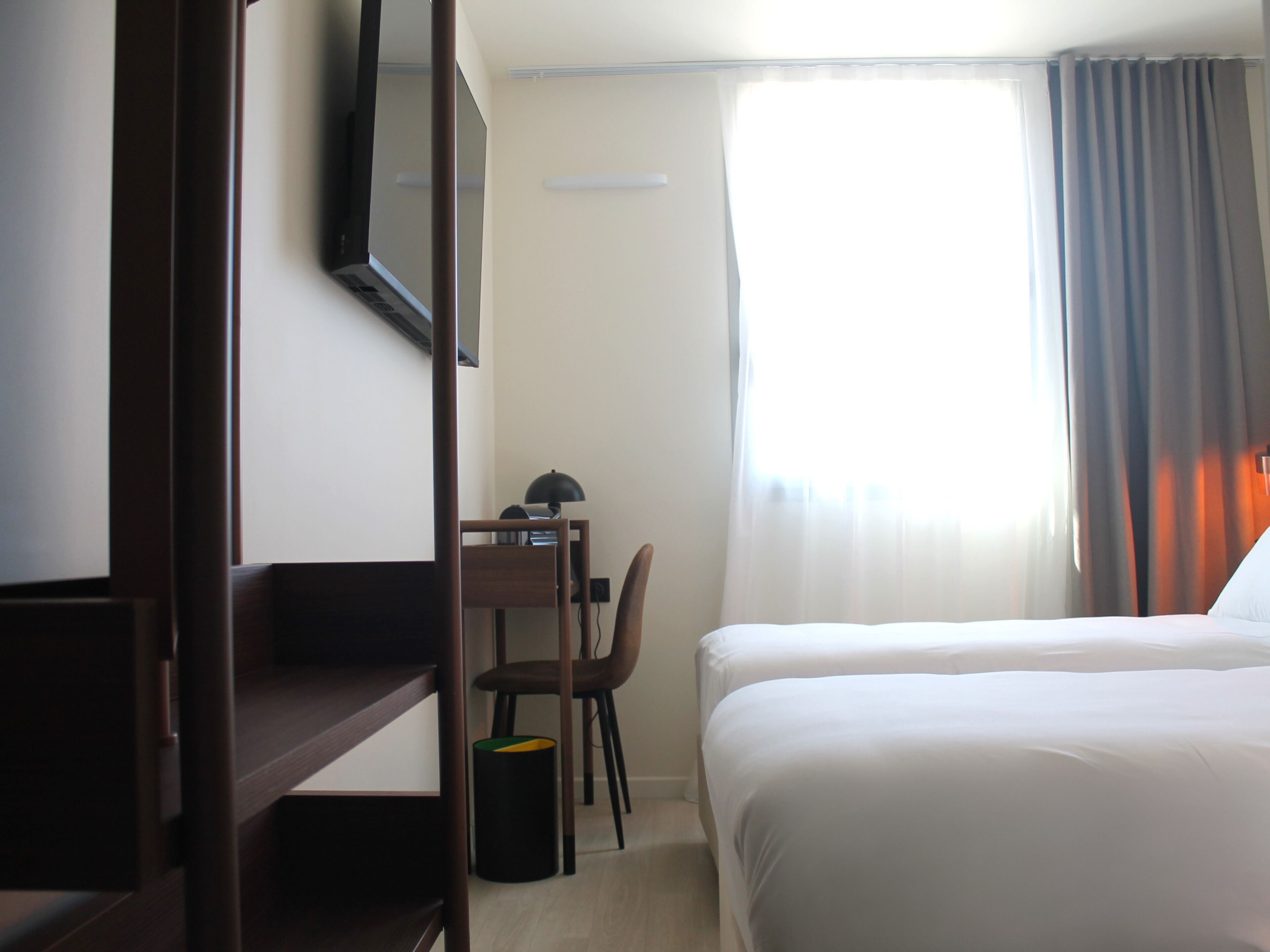 Una habitación de hotel con cama, escritorio con silla, televisor montado en la pared y ventana con cortinas.
