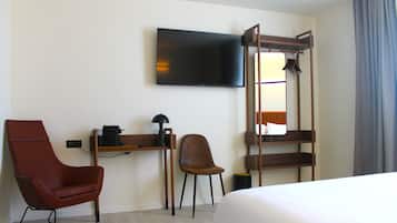 A hotel room with a bed, a TV, a red armchair, a brown chair, a desk with a coffee machine, and a mirror.