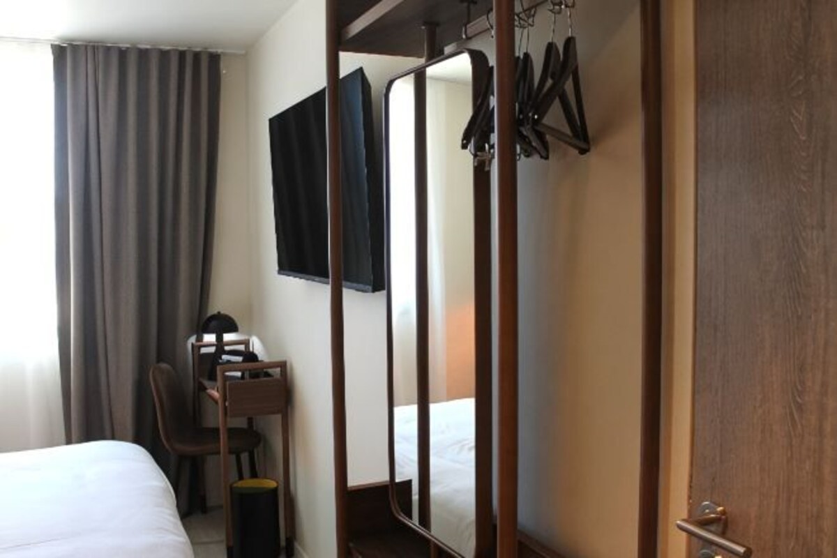 A hotel room with a bed, a desk with a chair, a mirror, and a coat rack.