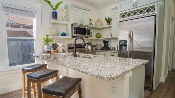 A modern kitchen with white cabinetry, a stainless steel refrigerator, a granite countertop, and two bar stools. - Brand New Craftsman 2BR/2BA Home in Denver Heights (San Antonio)