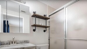 Une salle de bain avec un lavabo en marbre, des toilettes, une douche en verre et un miroir avec une étagère au-dessus.