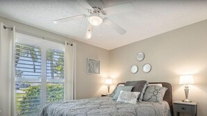 Une chambre à coucher comprenant un lit, deux tables de chevet avec des lampes, un ventilateur de plafond, une fenêtre avec des stores et un mur orné de trois assiettes décoratives.