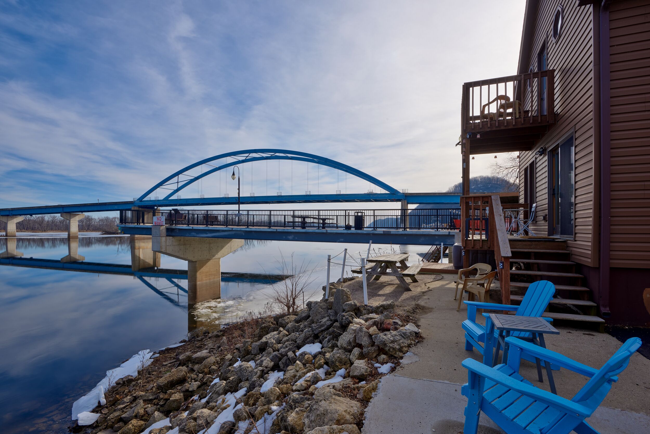 A bridge over a calm body of water, a wooden building with a balcony, and outdoor seating by the water.