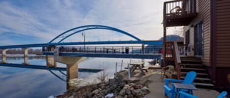 A bridge over a calm body of water, a wooden building with a balcony, and outdoor seating by the water.