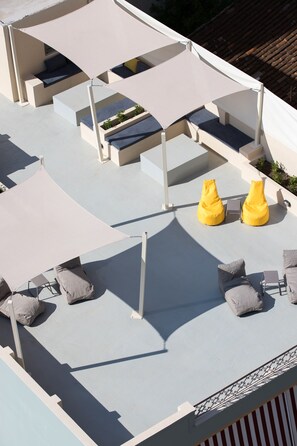 Una terraza en la azotea con zonas de estar, incluyendo sillones de mimbre amarillos y sofás grises, y un dosel blanco que proporciona sombra.