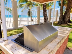 Un espace barbecue extérieur doté d’un couvercle en métal, entouré d’une clôture blanche, de palmiers et offrant une vue sur la plage.
