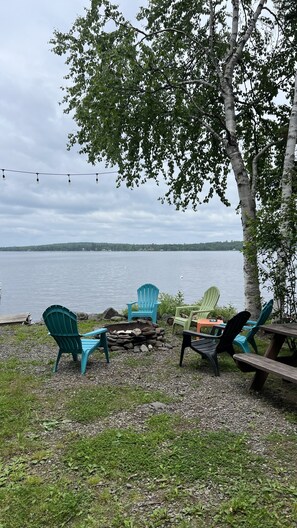 Un área de estar junto al lago con sillas Adirondack y una mesa de picnic, con vista a un lago tranquilo y una orilla lejana.