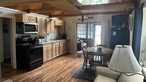 A cozy kitchen with wooden cabinets, a microwave, and a stove. There's a dining area with a table and chairs, and a lamp on the table. - Cozy escape in Portage Lake. Sleeps up to ten people!  (Portage Lake)