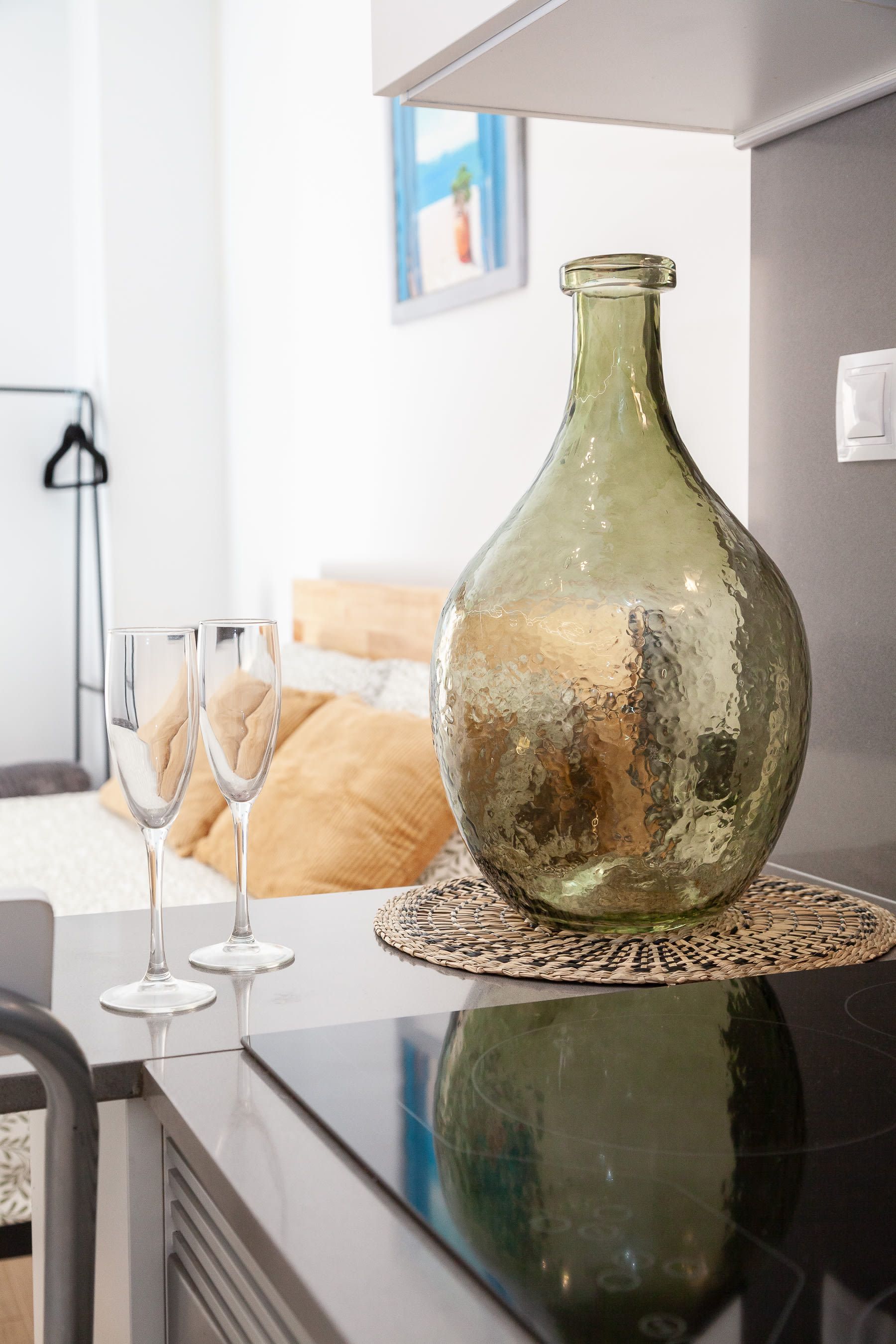 Ein Zimmer mit einer großen grünen Vase auf einer gewebten Unterlage, zwei leere Weingläser auf einer spiegelnden Oberfläche, einem Bett mit einer beigefarbenen Decke und einem gerahmten Bild an der Wand.