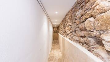 A narrow corridor with a stone wall on one side and a plain wall on the other, leading to a room with a stone floor.