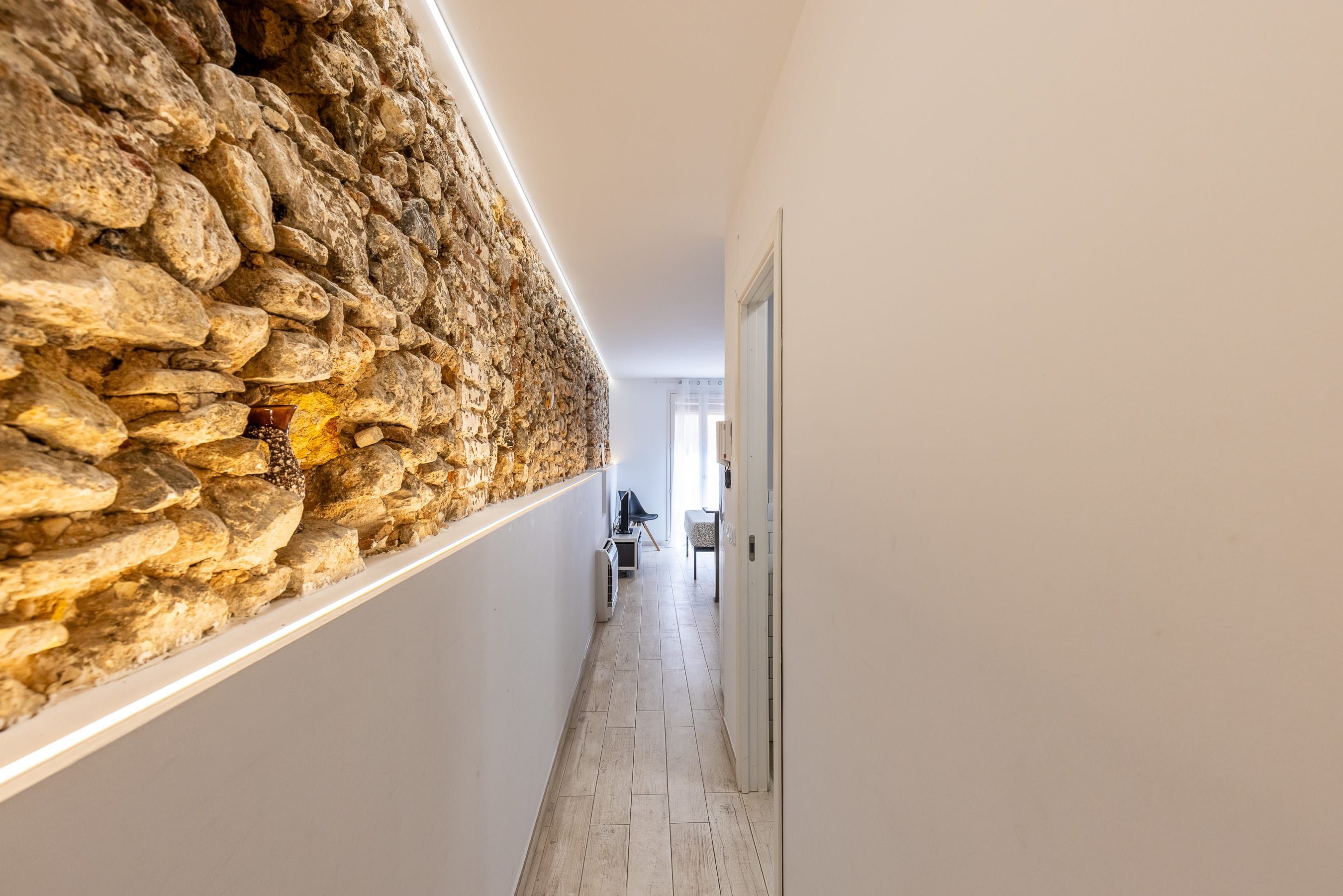 Um corredor com uma parede de pedra de um lado e uma parede branca do outro, levando a um quarto com uma cama e uma cadeira.