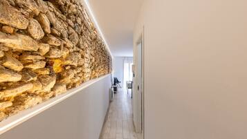 A hallway with a stone wall on one side and a white wall on the other, leading to a room with a bed and a chair.