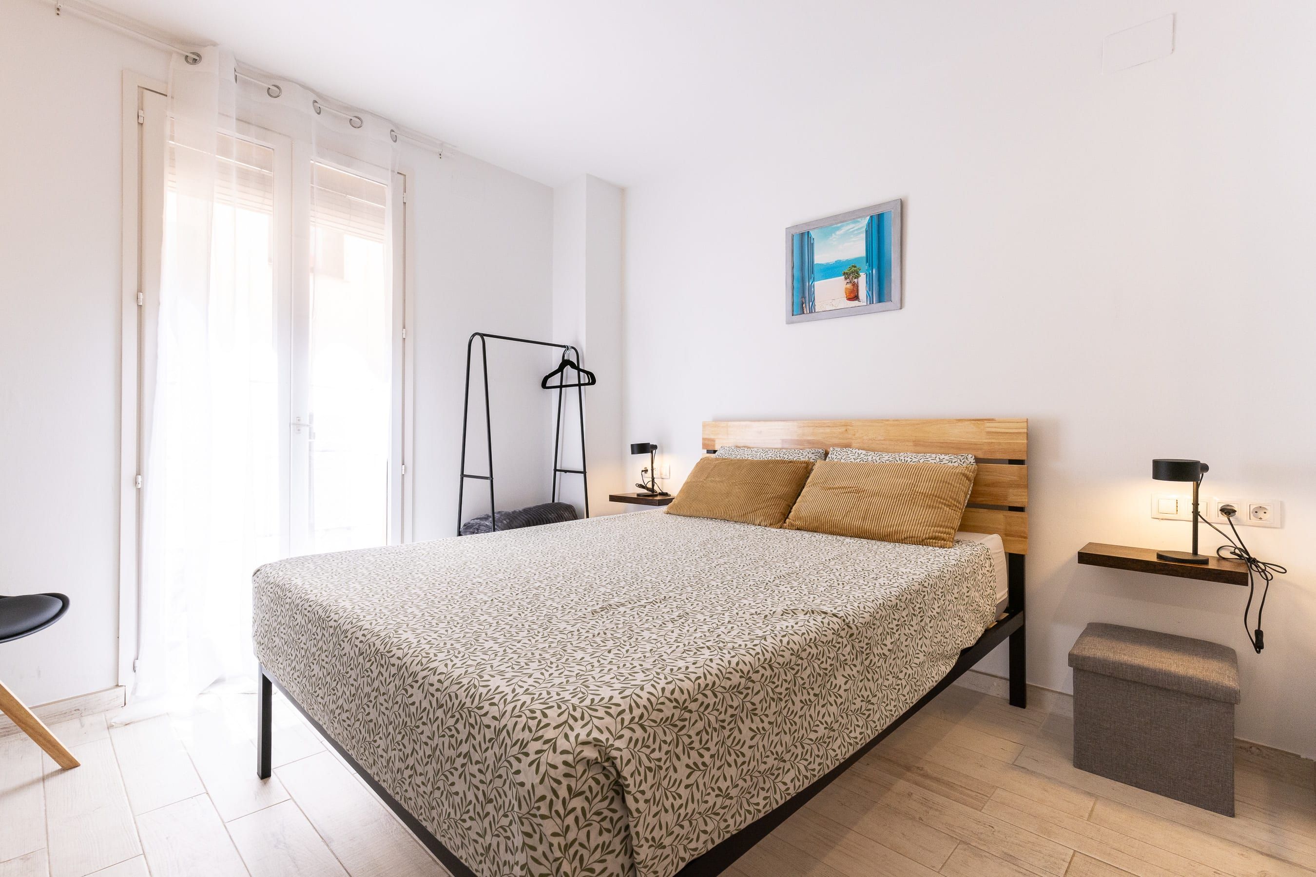 A bedroom with a bed, a bedside table, a nightstand, a hanging light, a picture on the wall, and a window with curtains.