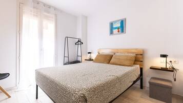 A bedroom with a bed, a bedside table, a nightstand, a hanging light, a picture on the wall, and a window with curtains.