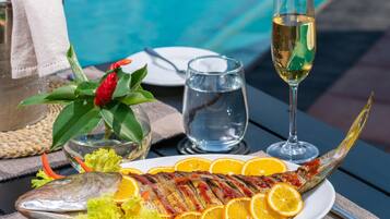 A dining table with a fish dish, a glass of wine, and a bottle of champagne by a pool.