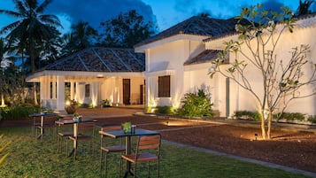 A resort with a covered outdoor seating area, palm trees, and a white building with a tiled roof.
