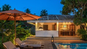 Zona junto a la piscina con sillones reclinables, un paraguas y una barra, con una cocina a la vista al fondo.
