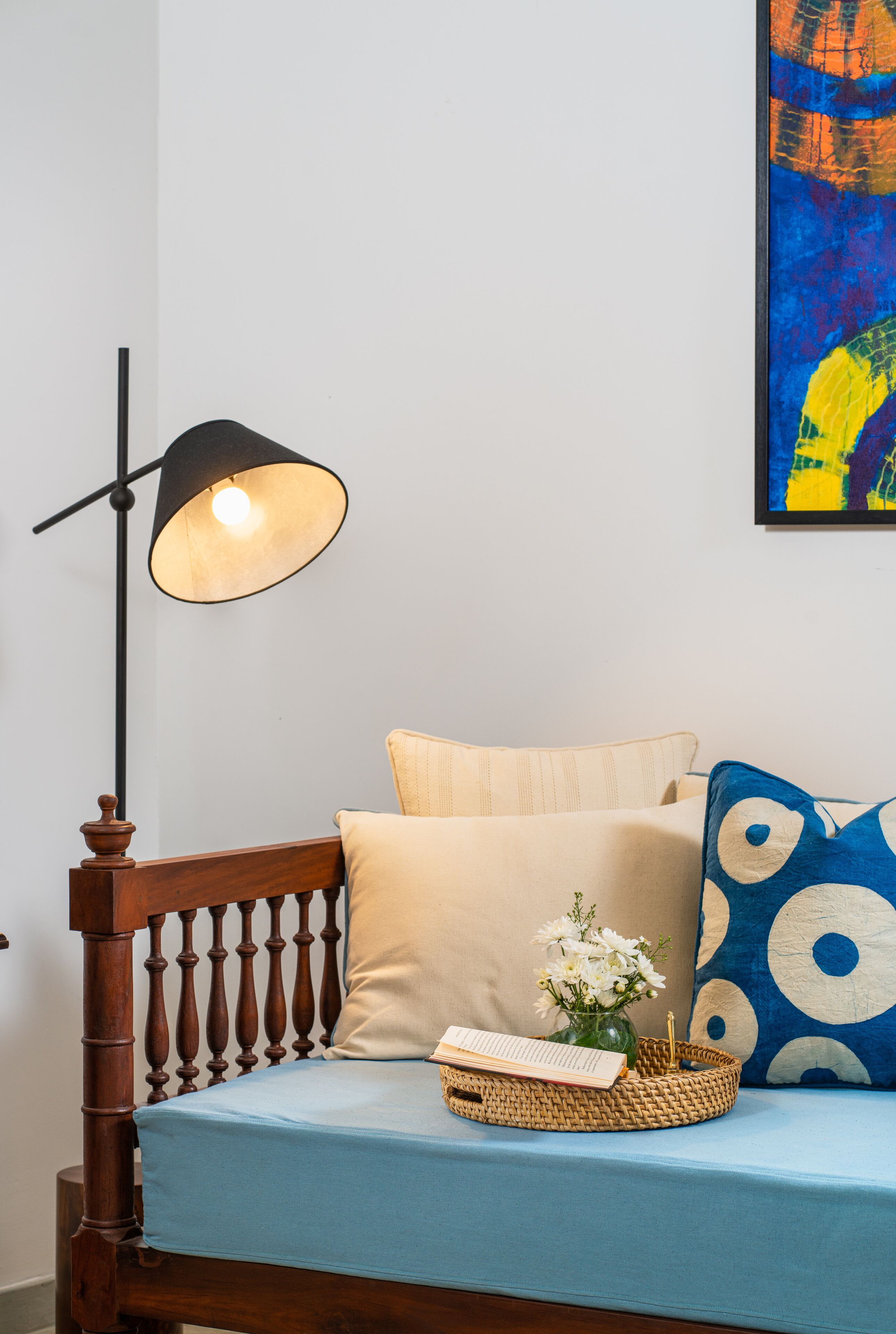Una cama con sábana azul, almohada color beige, almohada azul con diseño circular, un jarrón pequeño con flores y una bandeja tejida.