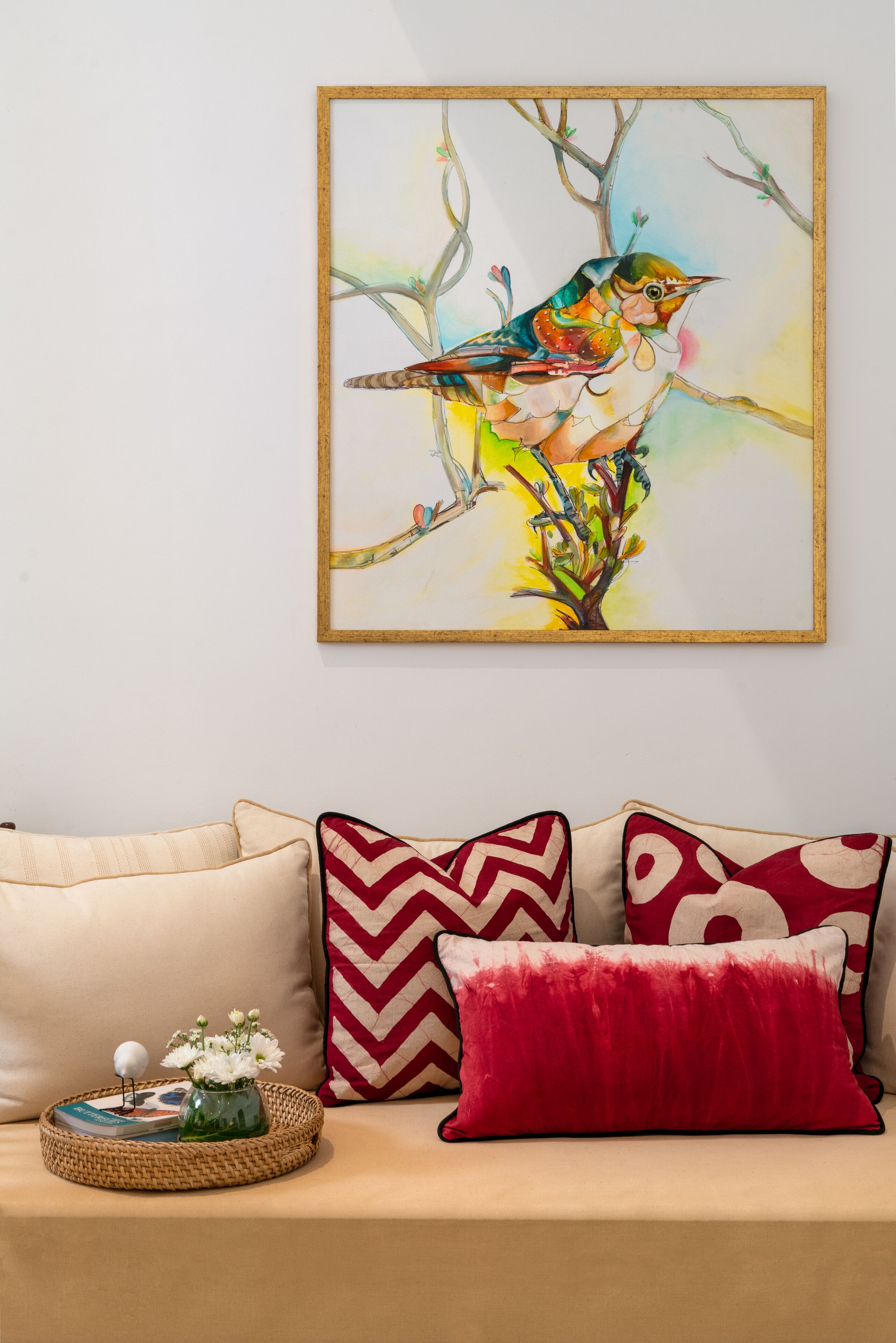 Una obra de arte enmarco que muestra un pájaro en una rama, un sofá color beige con cojines estampados, y una bandeja con un florero y libros.