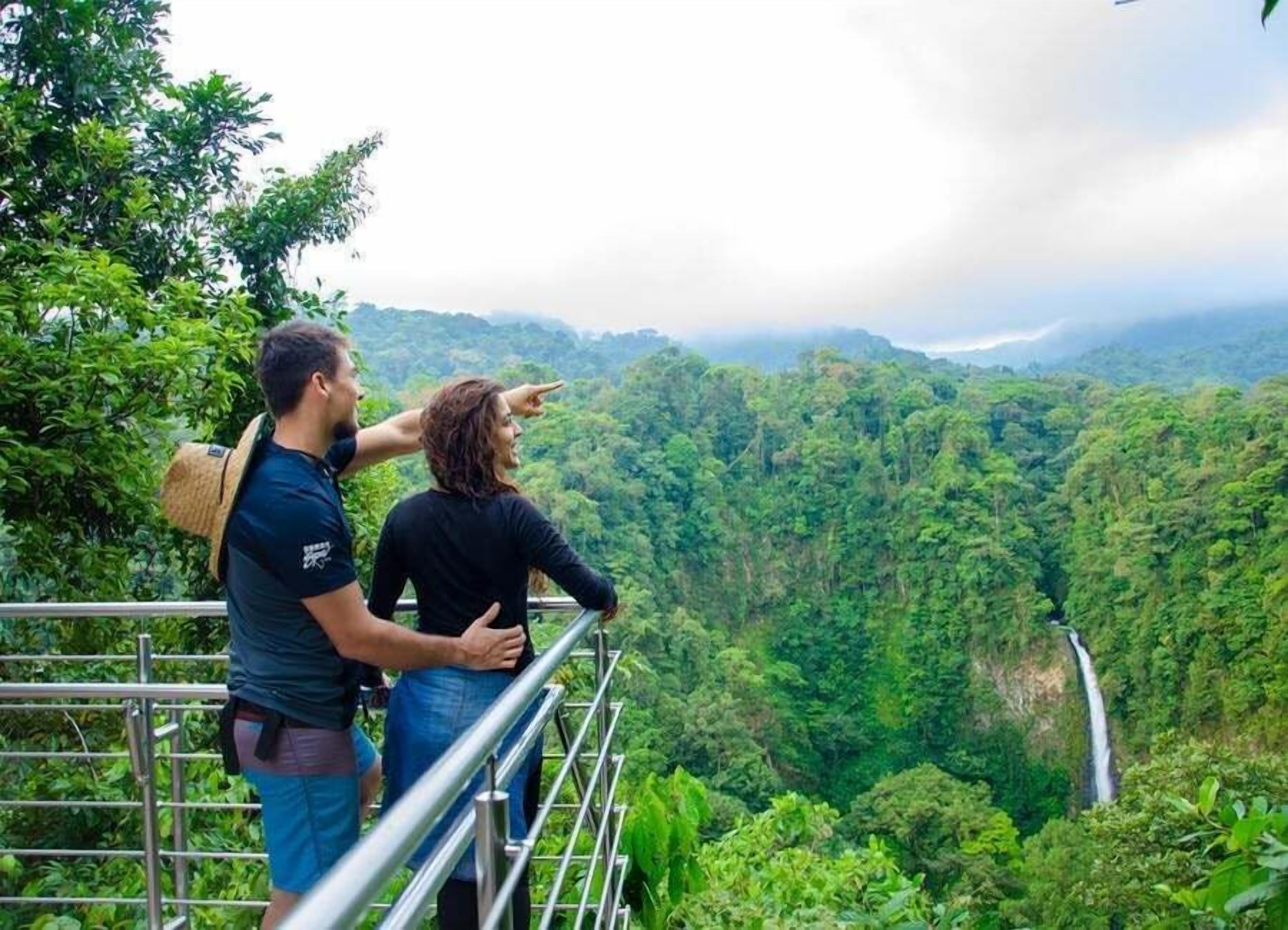 A couple standing on a metal railing overlooking a lush green valley with a waterfall.