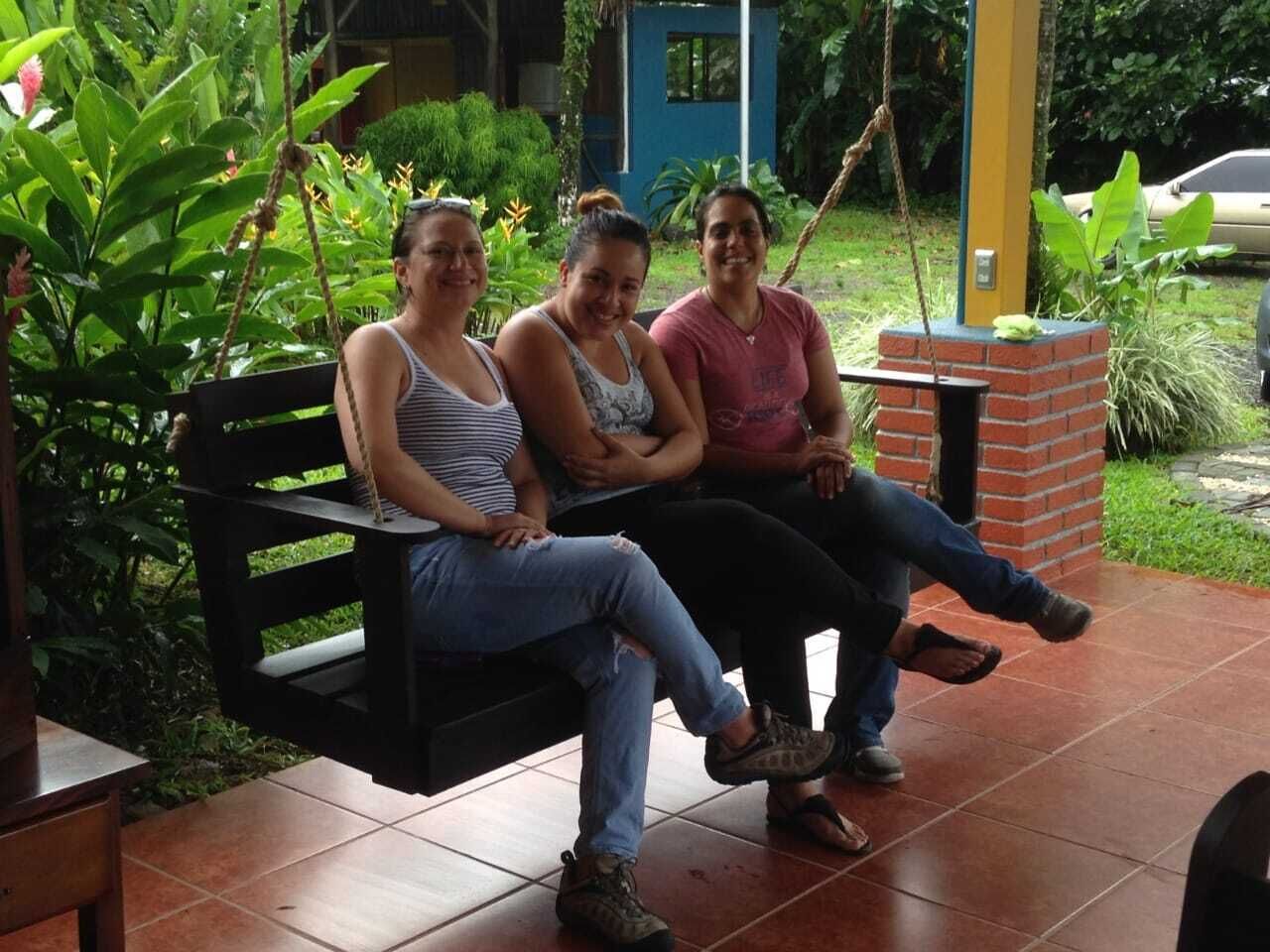 Trois personnes sont assises sur une balançoire dans un jardin, avec une structure en briques et de la verdure en arrière-plan.
