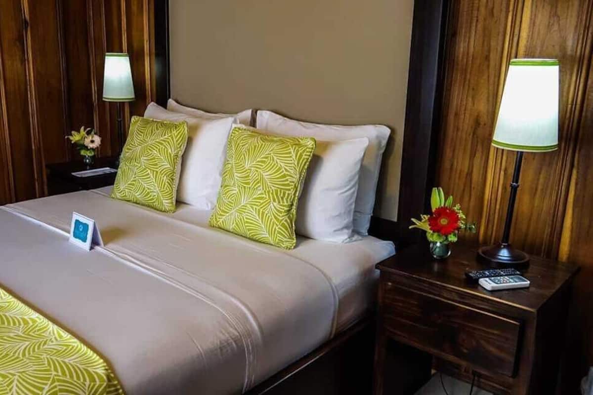 A hotel room with a bed, two bedside tables, a nightstand, a lamp, a remote control, and a small vase with flowers.