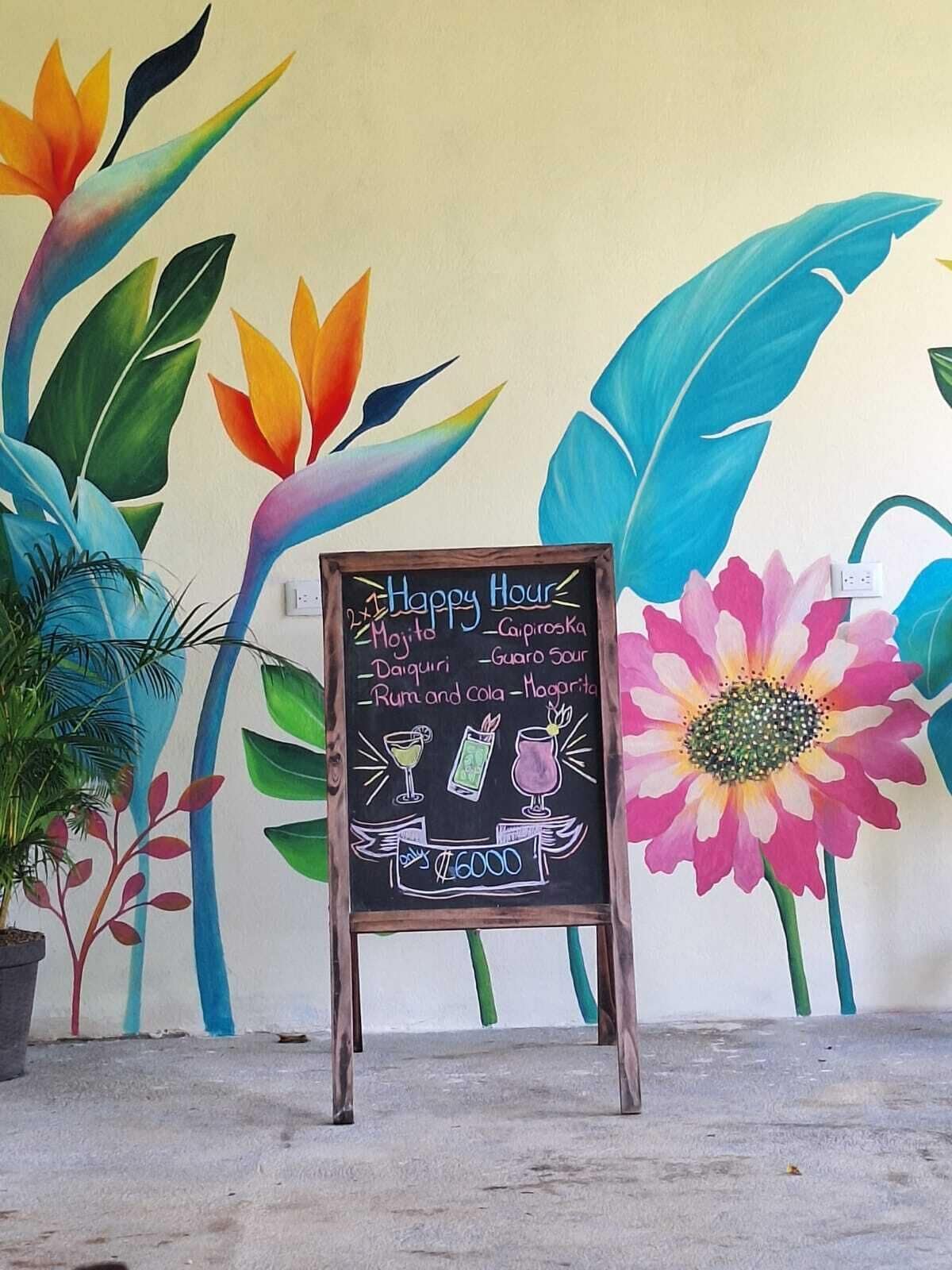 Un letrero de pizarra con un menú que enumera bebidas y precios, frente a un muro con murales florales coloridos.