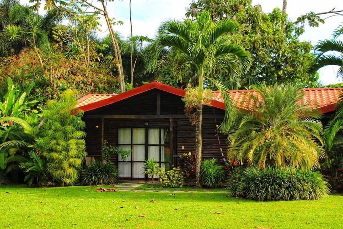 A wooden cabin with a red roof, surrounded by lush tropical vegetation.